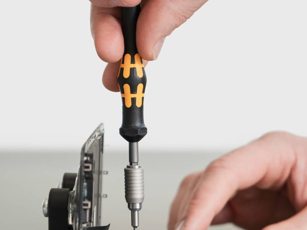A phone held in a repair jig with the back removed, while a precision screwdriver is used on an internal screw
